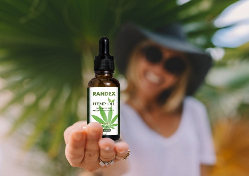 Woman holding a bottle of Randex Organic Hemp Oil 5000MG, made in the USA, in a sunny outdoor setting with palm trees.